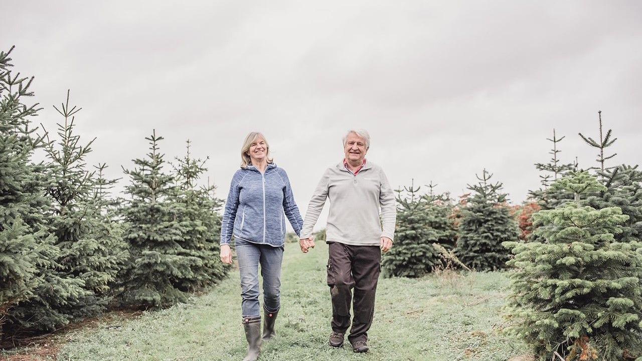 Alastair and Diane stood arm in arm in a field. There are Christmas trees around them.