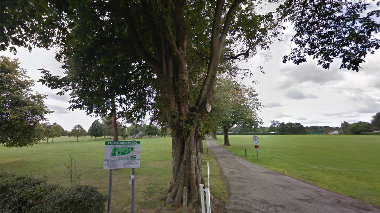 A wide view of Queens Park in Caterham, Surrey, showing trees in full leaf and lawns with a concrete path running between them.