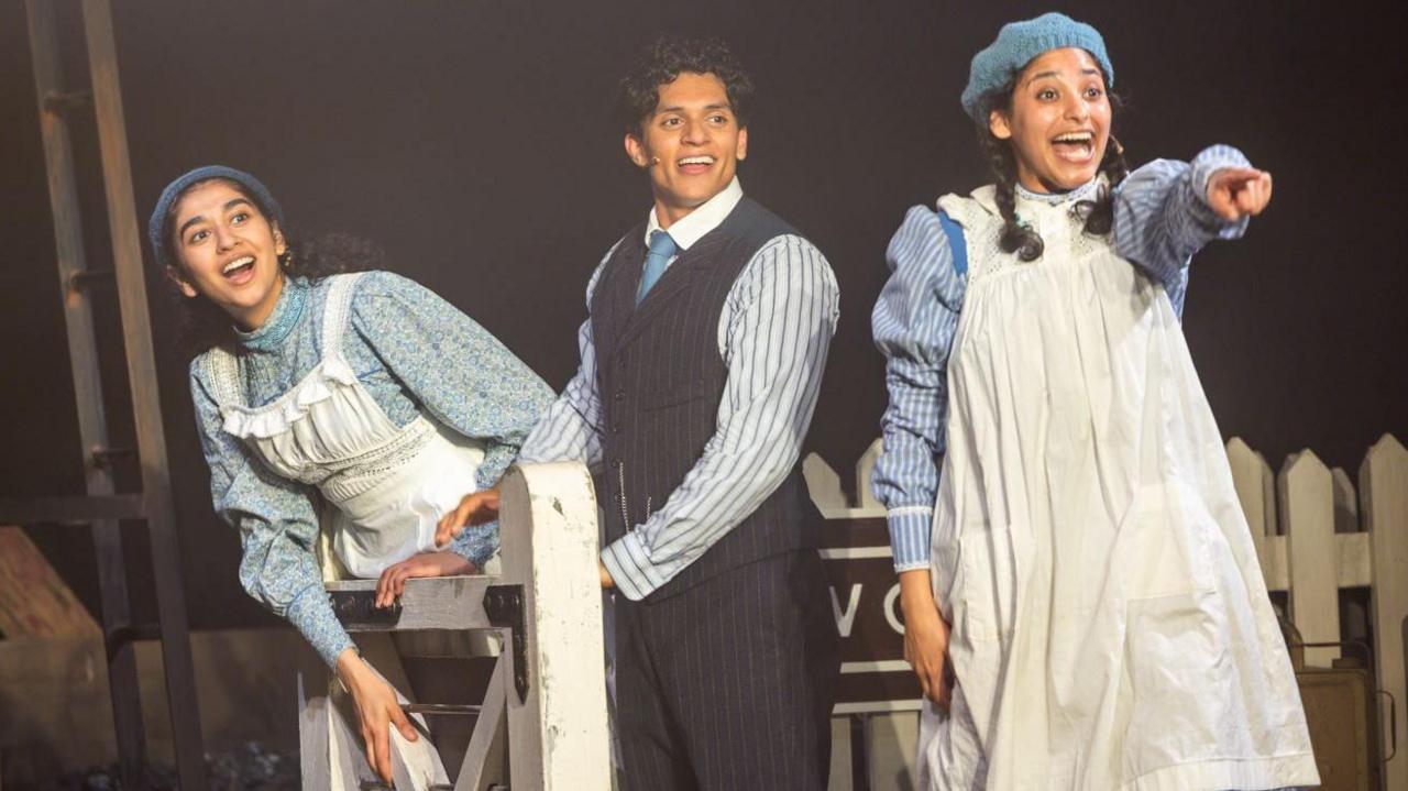 Three Railway Children actors stand on a platform and look to their left. One is pointing and all are smiling. 
