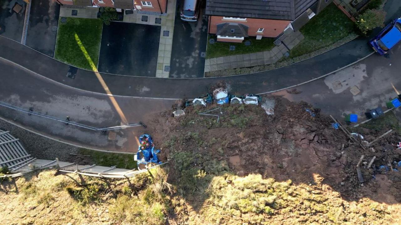 Aerial view of the landslide, showing  soil covering the road and temporary fencing on the left next to the bank