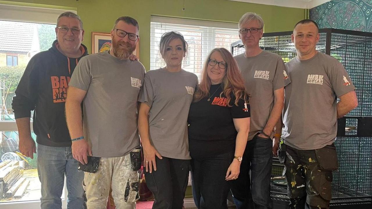 A group of four builders standing with Simon Taylor and Debi Richens. Simon is tall and slim with grey hair and glasses. Debi is shorter with light pink hair. The tradespeople are wearing paint-splattered clothes.