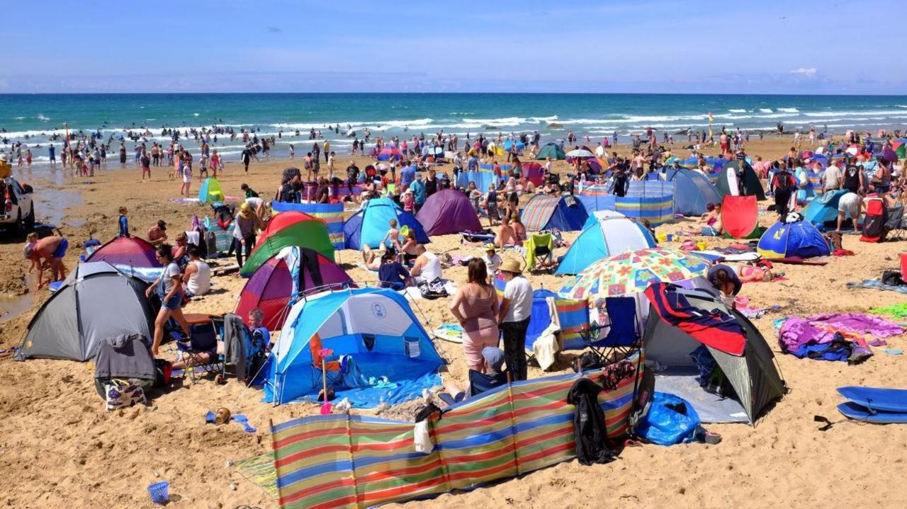 Dozens of people gathered on the beach at Watergate Bay in Newquay, Cornwall, during a sunny day. Several tents and beach wind screens have been set up on the sand. People are heading out into the sea on a sunny day.