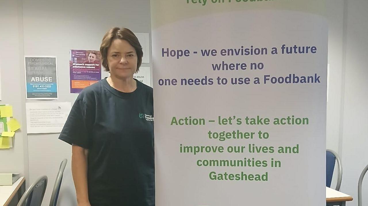 Diane Kindred standing next to a Gateshead Foodbank banner. She has dark hair and is wearing a dark blue T-shirt. The banner includes a number of action points such as "Hope - we envision a future where no one needs to use a foodbank".