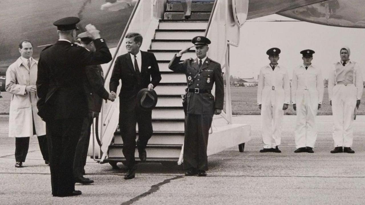 A black and white photograph of the Boeing Air Force One plane on a runway. President Kennedy is at the bottom of the stairs shaking hands with one of the waiting party. Several military personnel are nearby standing to attention.