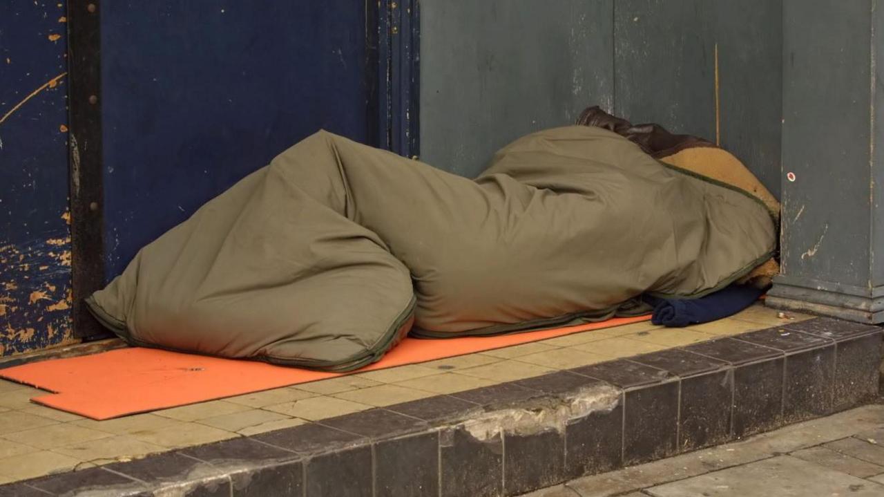 A khaki sleeping bag on a step outside a building with a blue door with someone appearing to be in it. An orange cardboard sheet is underneath the sleeping bag