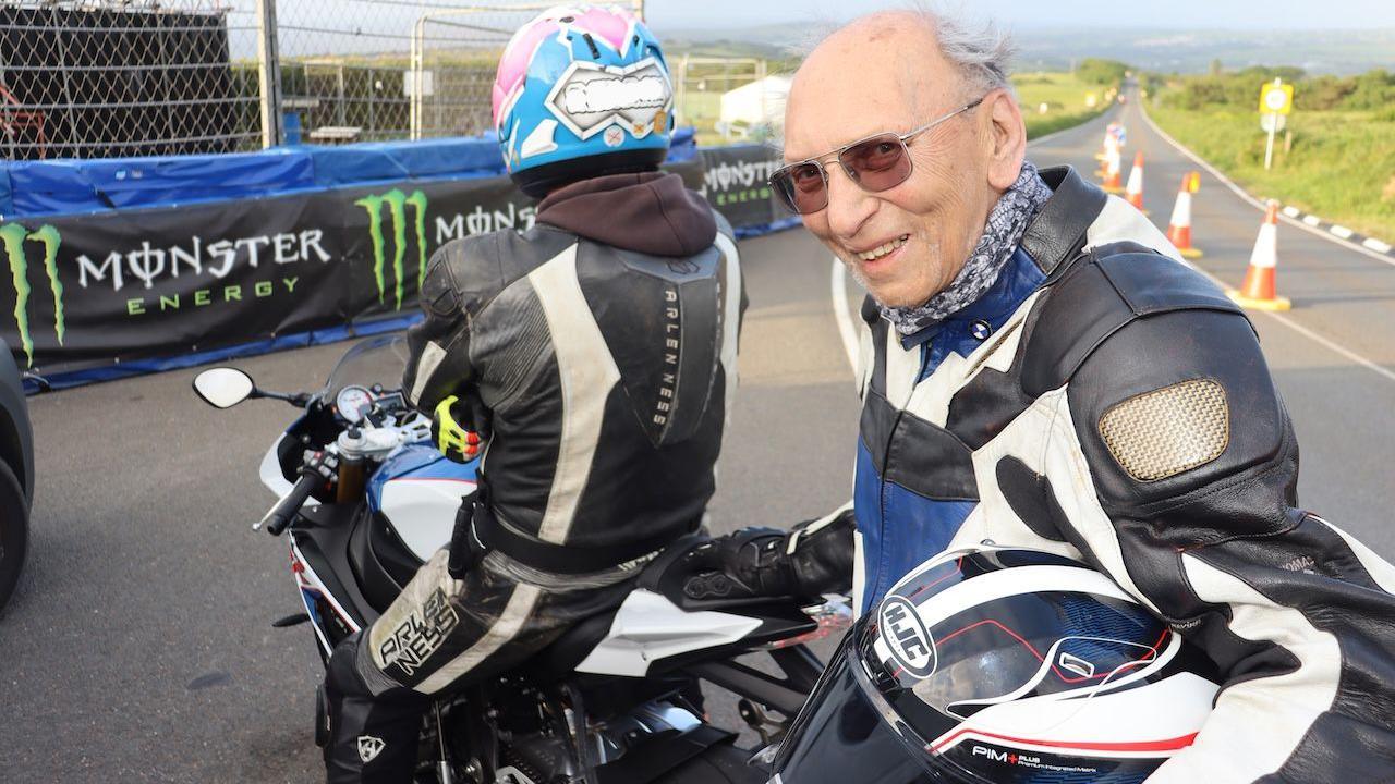 Ron, an elderly man smiles in leathers he holds a helmet and wears sunglasses and turns around on the course.