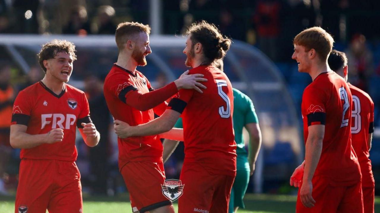 Four FC Isle of Man players celebrate a goal. They are wearing red shirts and shorts which have black cuffs.
