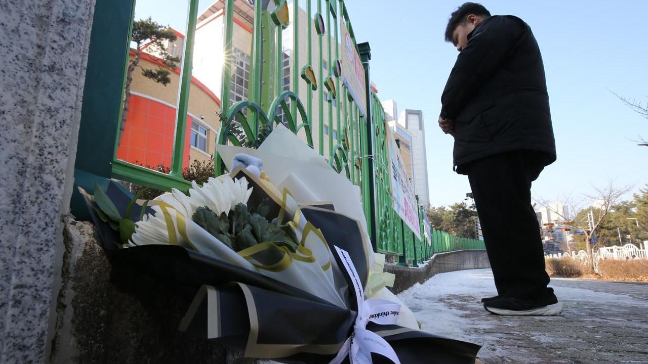 A man standing with his hands crossed in front of a school gate. Bouquets of flowers lay on the ground beside him.