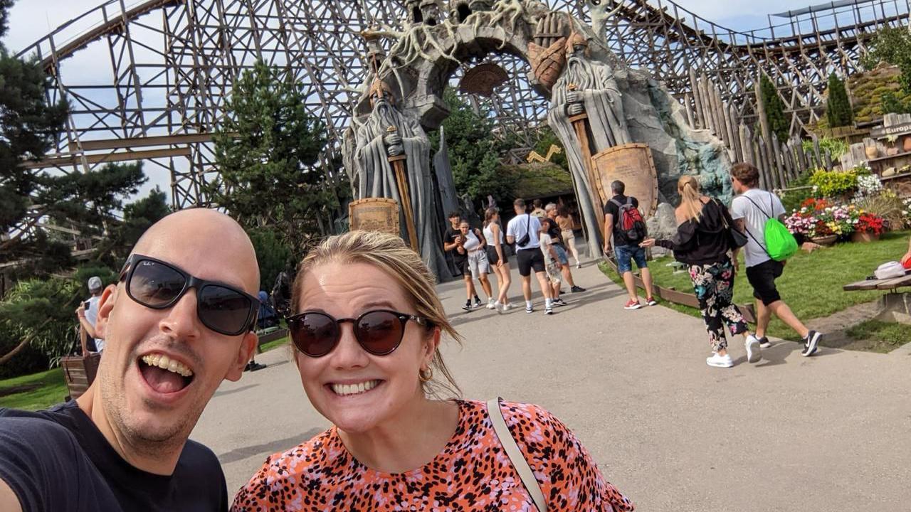 Dean Stokes - a man with no hair smiles into the camera. He is wearing sunglasses and next to a woman, also wearing sunglasses. She is smiling, wearing a pink and black dress and has blonde hair.