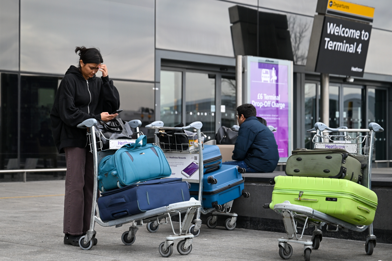 A man and a women with suitcases piled on trolleys outside Terminal 4. The woman is looking at a phone and the man is sitting down on a wall