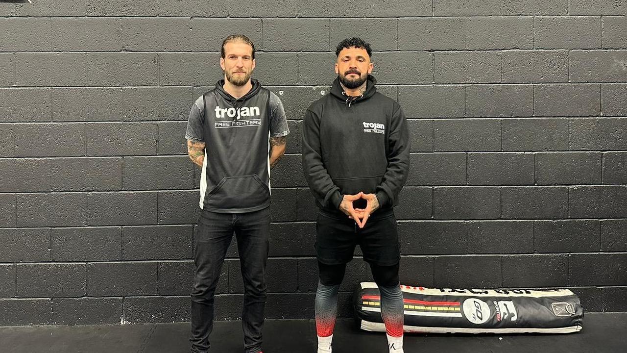 Mark McLelland and Mario Saeed standing in front of a stone wall painted black, and a punching bag on the floor 