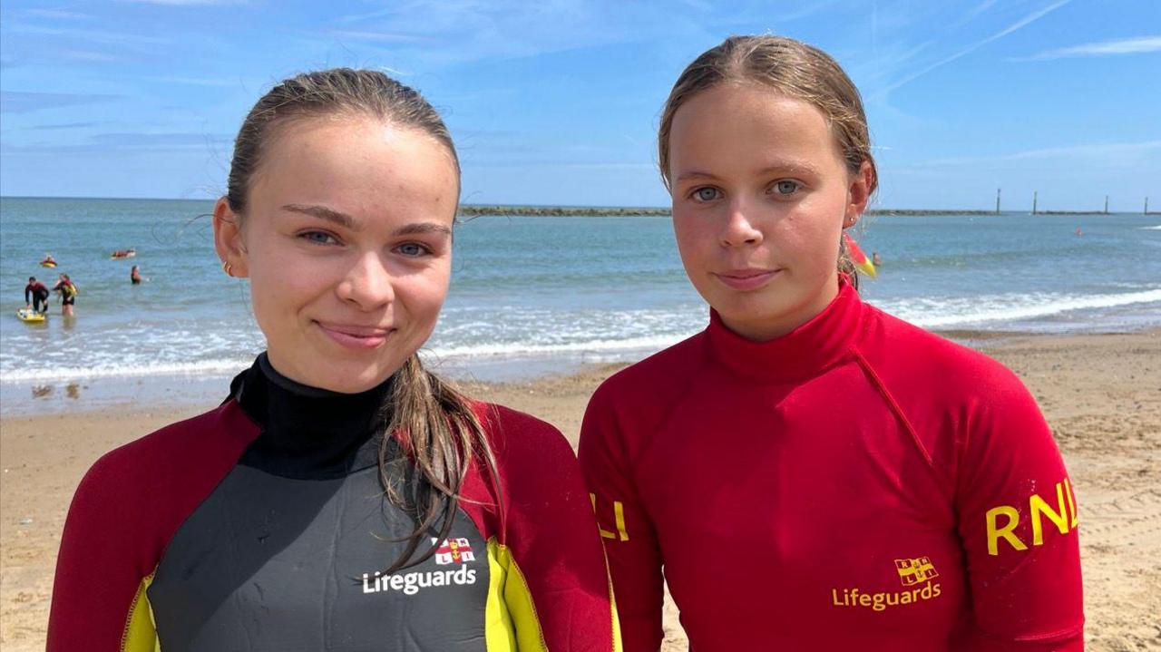 Phoebe Claybourne and Emily both standing next to each other on the beach at Sea Palling in Norfolk. They are both looking directly at the camera and are smiling. Behind them is the sea and a blue sky.