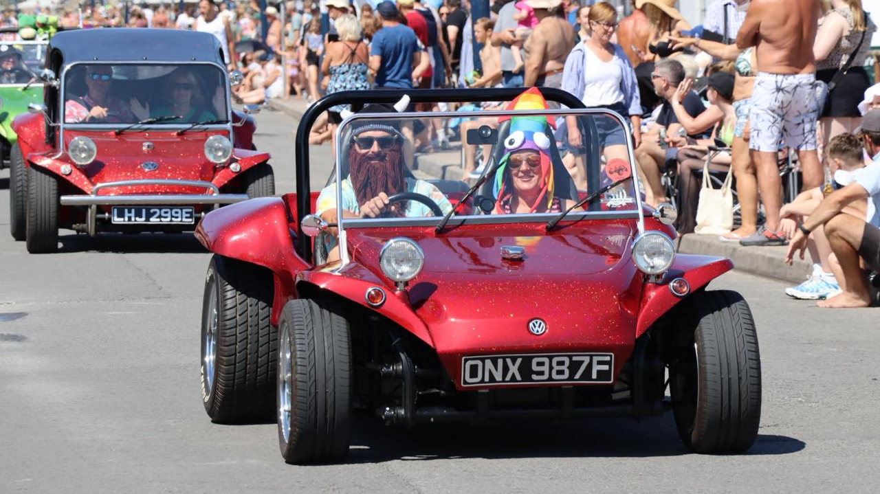 Two people sat in a red VW car, which is sat low to the ground, driving through the carnival.