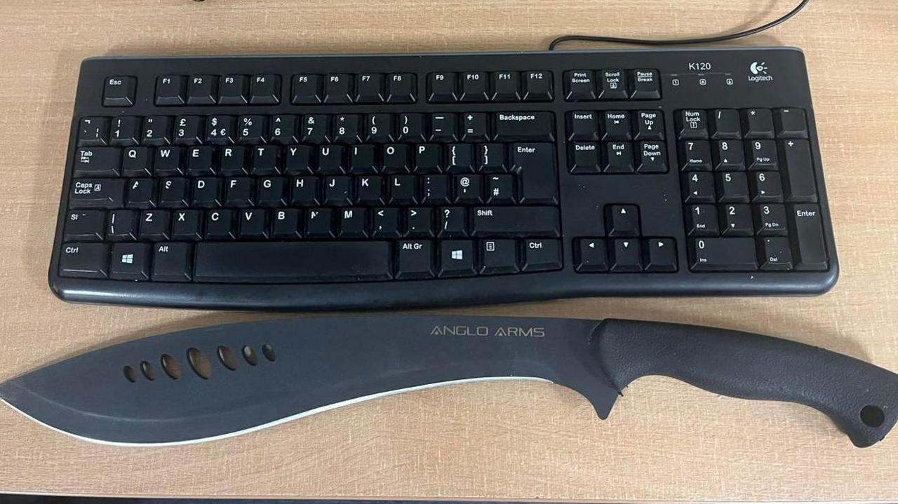 A machete with a black handle and "Anglo Arms" branding on a light-coloured wood desk, next to a black computer keyboard. The machete is about two inches longer than the keyboard.