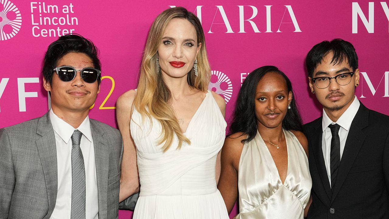 An image of Angelina Jolie wearing a white dress and three of her children on the red carpet standing in front of a pink board 