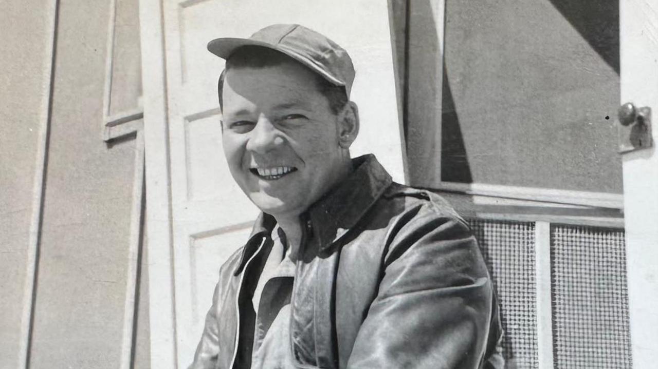 Joseph Gray wearing a leather jacket and soft cap on an angle, smiling. It is in black and white.