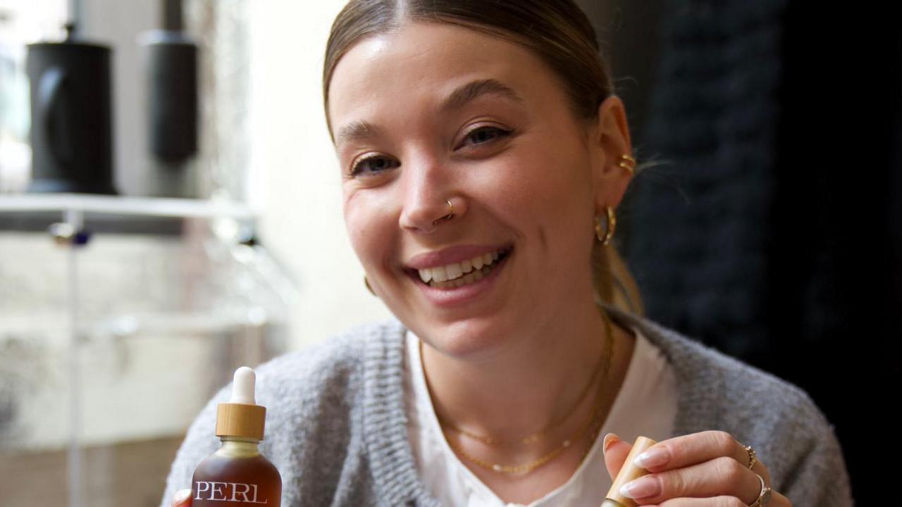 Isobel, wearing a white top and grey cardigan, smiles at the camera and is holding two of her beauty products