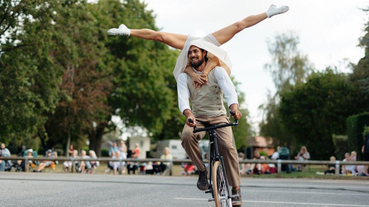 A man rides a black cycle with only one wheel visible, while a second performer balances upside down on his back, arms wrapped around the man's shoulders, legs in straddle splits in the air. The upside-down performer wears white shorts, boots, and what appears to be a skirt, which is draped around the cyclist's shoulders. The cyclist has dark hair and facial hair, and is wearing light brown trousers and shoes, a beige sleeveless jumper and a white shirt.