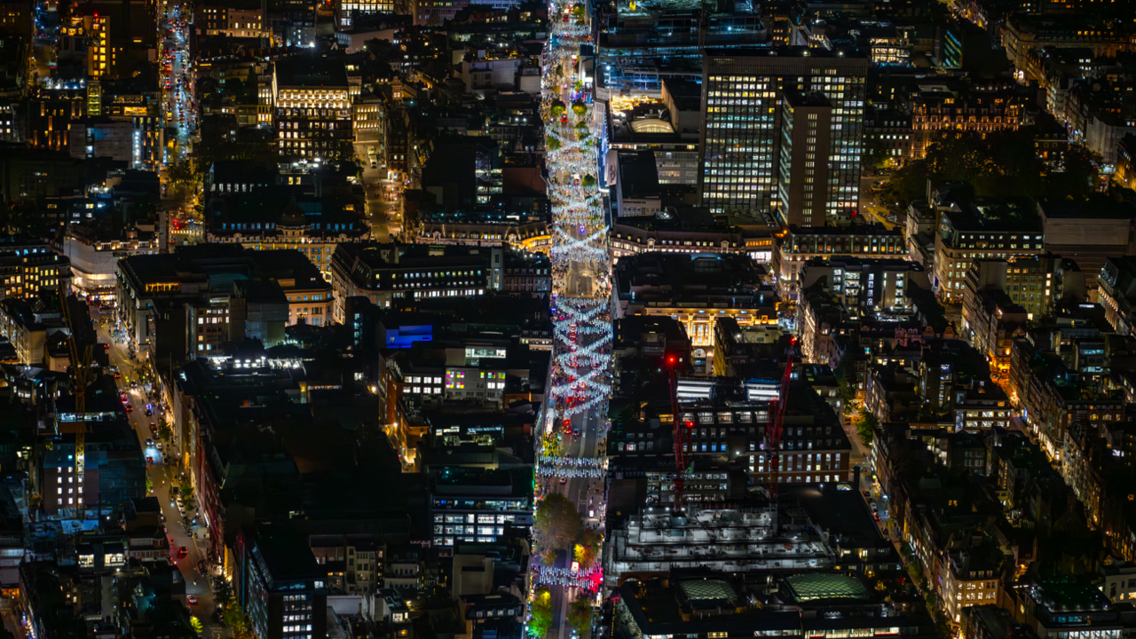 Wide aerial photograph taken at night showing white/blue Christmas lights hanging across Oxford Street with buildings with lights on in them either side of it