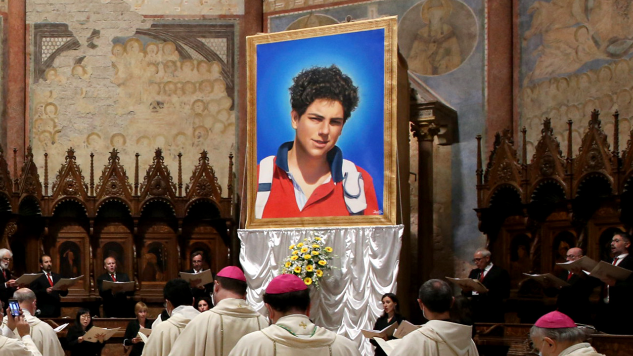 A portrait of Carlo Acutis on a platform in a cathedral. He is a teenage boy in a red jacket. There are people wearing Catholic religious attire and the cathedral has gothic architecture.