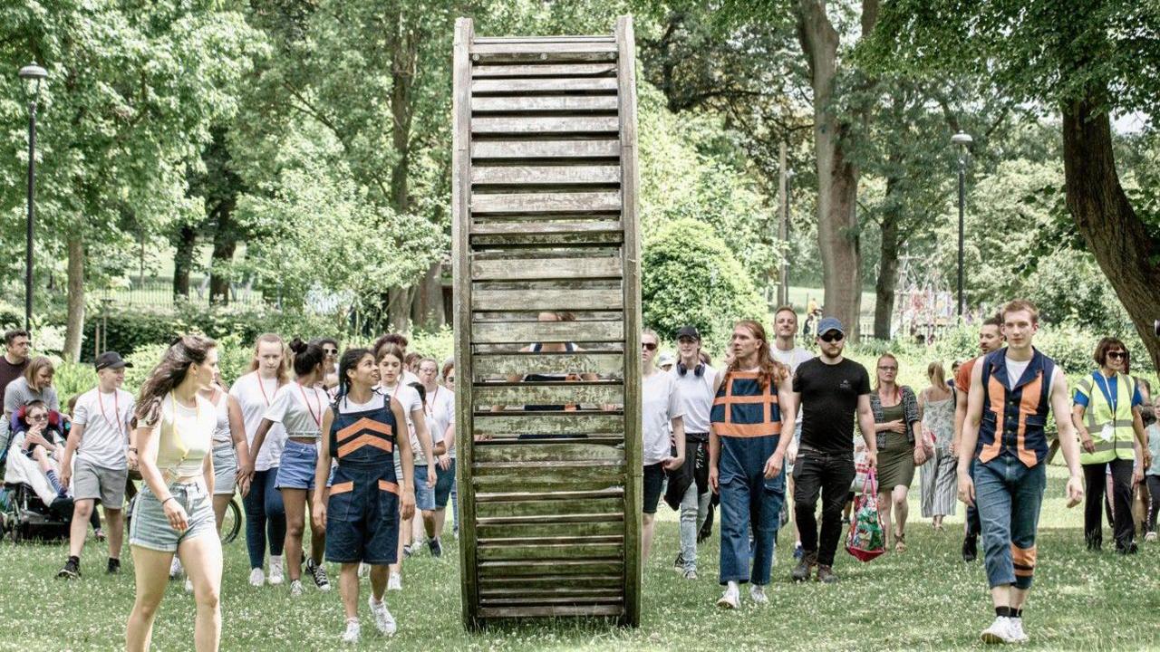 People of various ages walk with a large wooden wheel structure through green parkland, accompanied by three people wearing navy blue outfits with orange trim, and one person wearing a yellow hi-vis jacket.