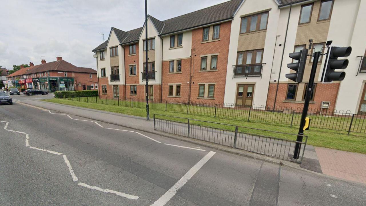 Knaresborough Road in Harrogate - a pedestrian crossing across a main road leading to modern flats in front, a parade of shops to the left (including Drake's Fish and Chips on the corner) and several cars on the road and parked near the shops