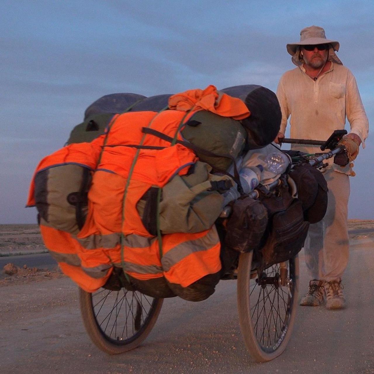 Karl Bushby wearing dusty clothes, sunglasses and a wide brimmed hat is pushing a cart, which contains his belongings covered in orange hi-vis fabric, along a dusty road.