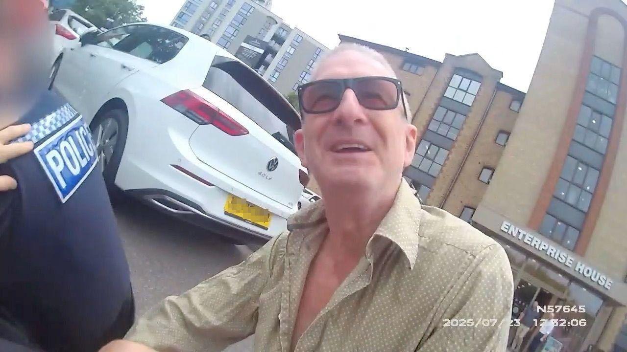 Body-worn camera image of a smiling Jeff Stewart squatting behind a policeman in a car park. He is smiling and is wearing sunglasses and a pale shirt with several buttons open at the chest.