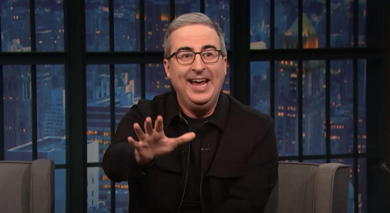 John Oliver sits on a talk show chair speaking to camera - he is animated and reaches his hand out, gesticulating. He wears black glasses and has short grey hair and dark eyebrows.