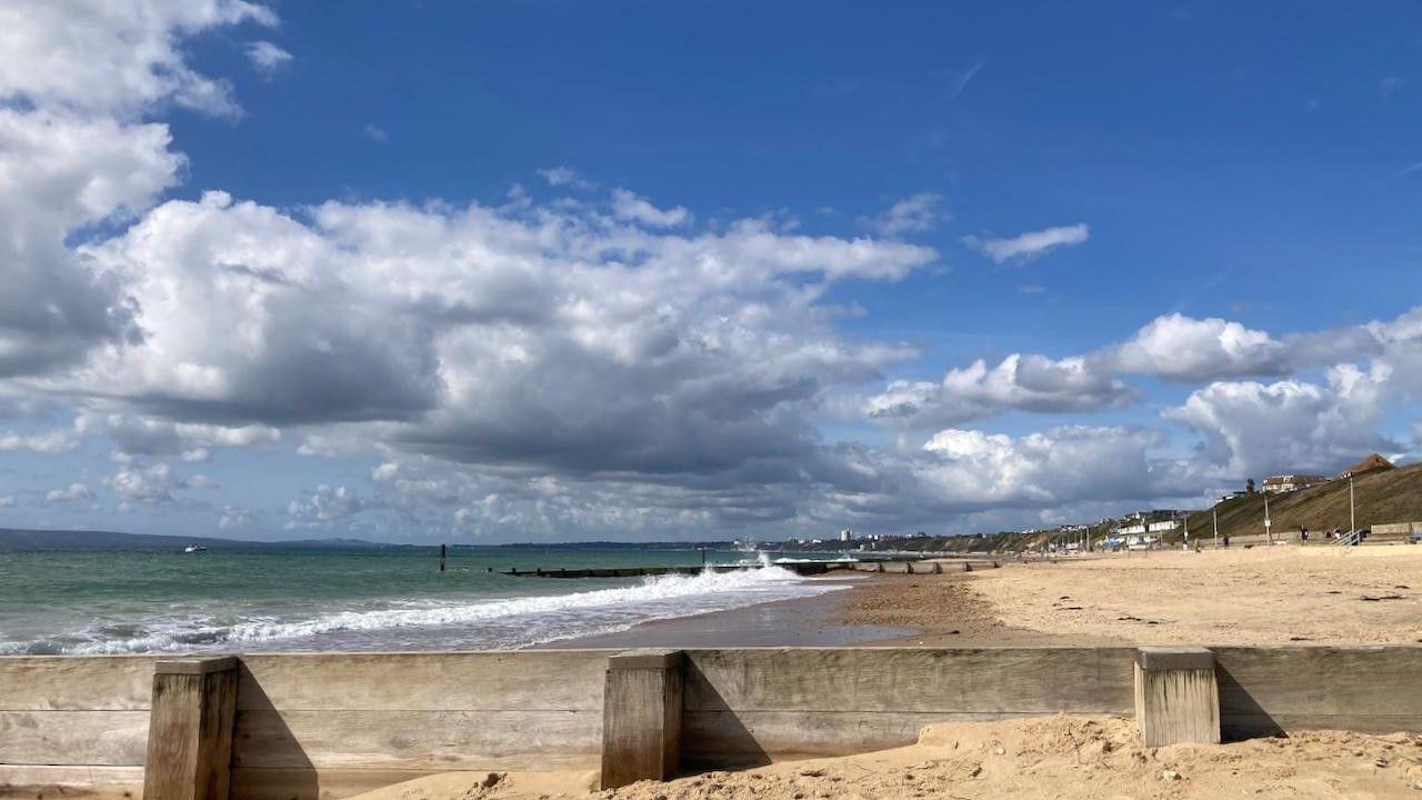 a sandy beach with waves crashing against wooden groynes, green sea, buildings in far distance, under a cloudy blue sky