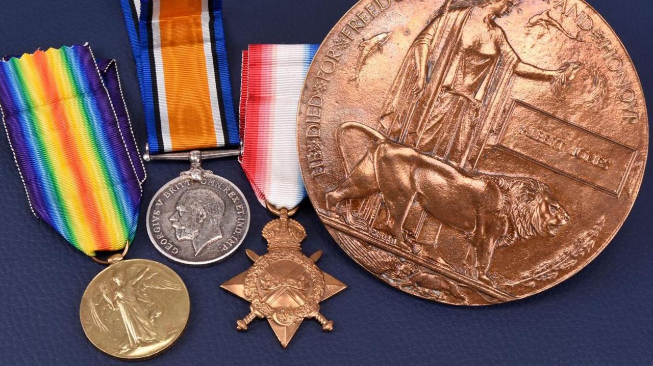 A collection of three metal medals on multi-coloured ribbons and a much larger circular metal coin all sit on a dark blue background.