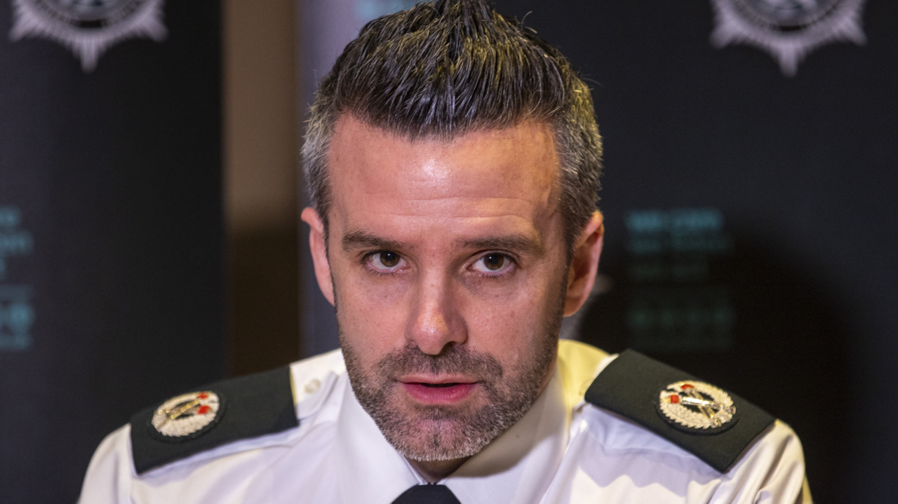 A police officer wearing a uniform - white shirt and black tie - looking into the camera. He has a short dark beard and dark hair.