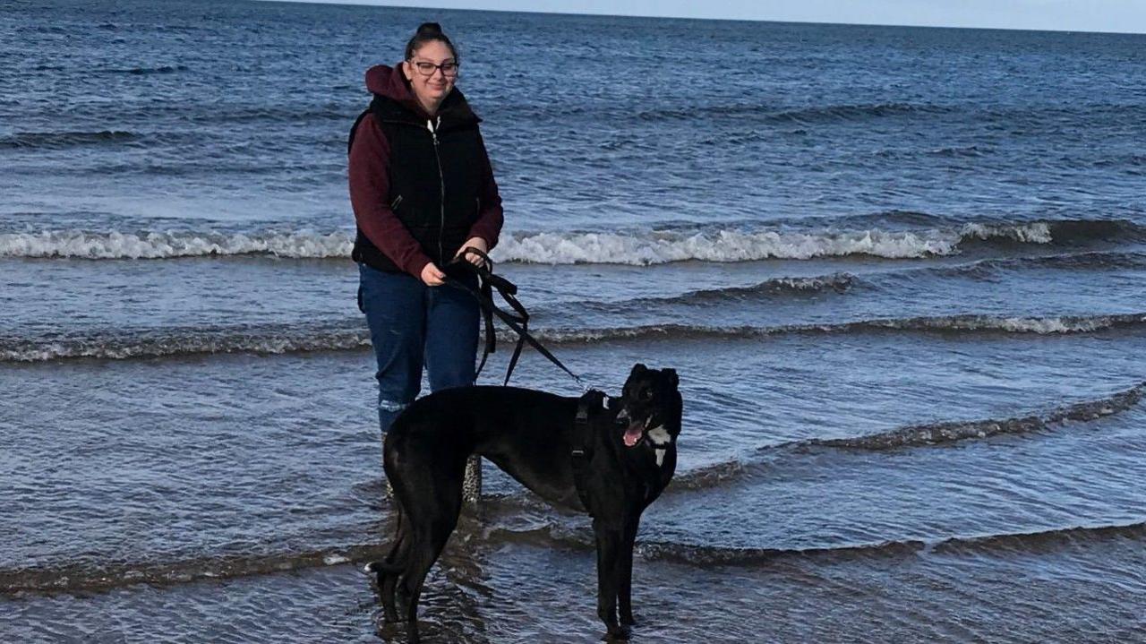 A photo of Lotte stood with her dog Tina, a black whippet, in the shallow waves of a beach. She has walking boots, blue jeans rolled up, and is wearing a dark gilet over a maroon hoodie.
