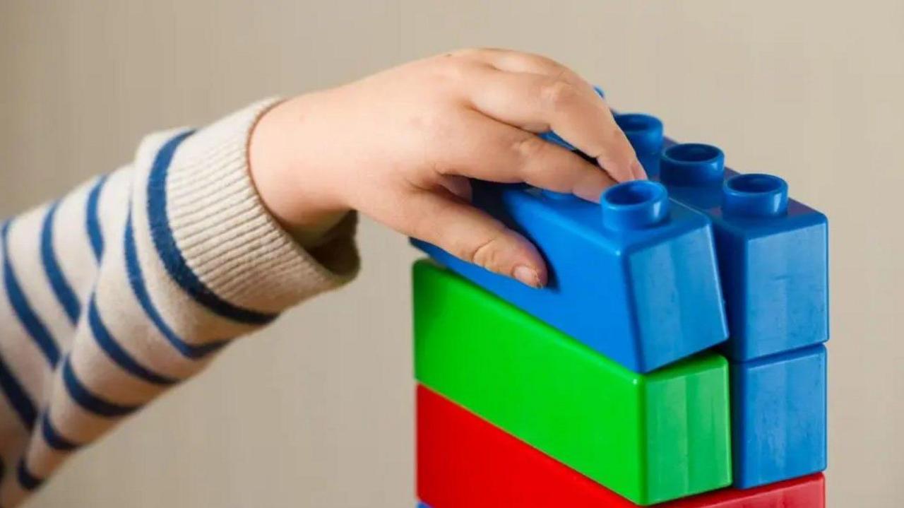 Toy bricks - some are blue, others are red and one is green - are on the right hand side of the photo. A hand is touching one blue brick on the top of the pile.