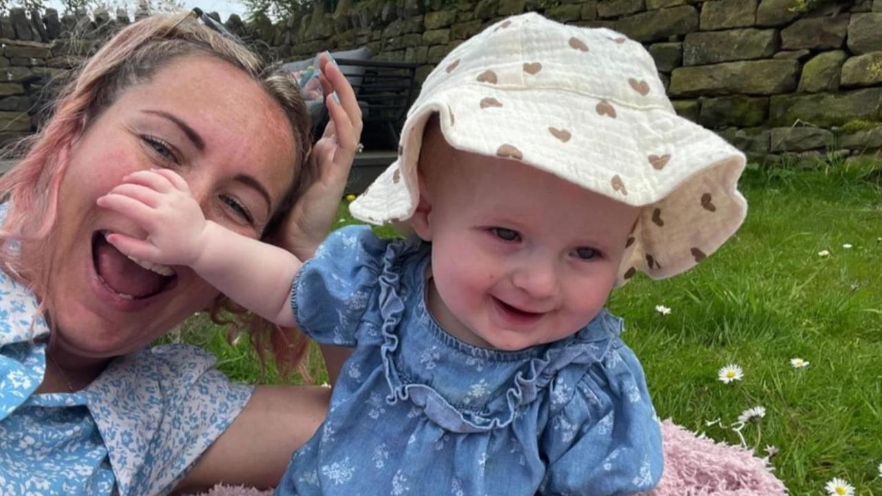 Penelope and her mum playing on grass. Penelope is wearing a blue dress and white floppy hat.