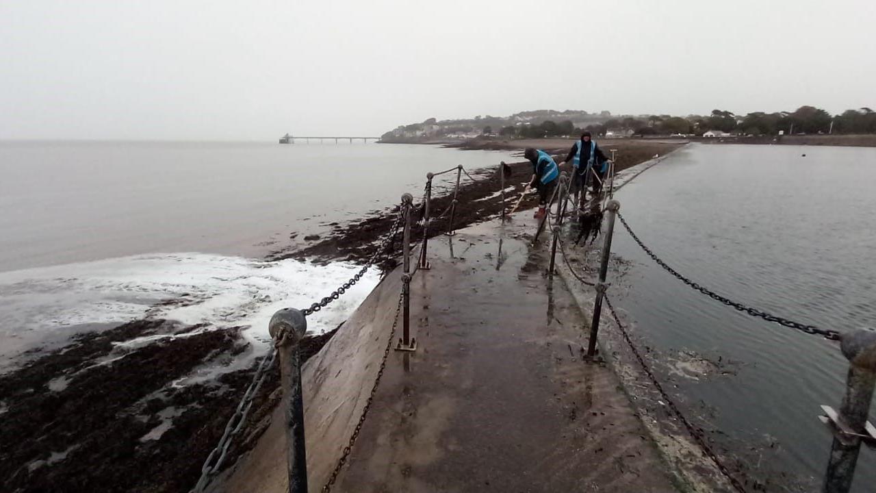 The wall between Clevedon Marine Lake and the sea next to it. People are on the wall cleaning it. They are wearing blue high vis vests.