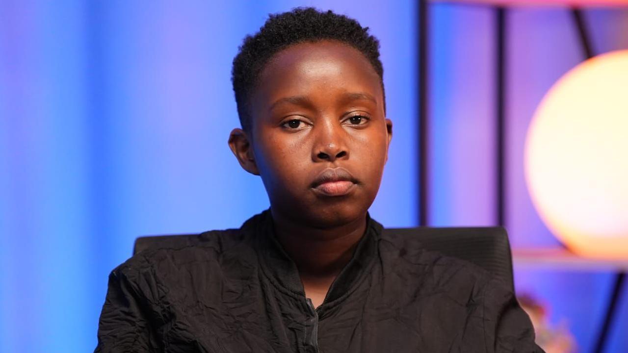 Esther Njoki stares at the camera. She's drressed in a black jacket in a purple room, with a bright, circular cream-coloured light in the background