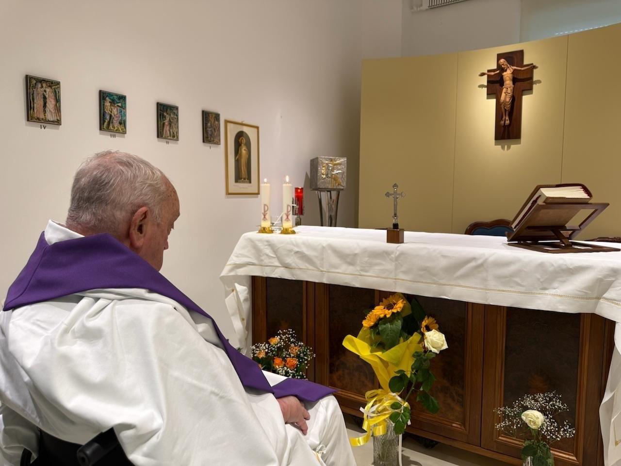 The pope sitting in a wheelchair looking at an altar in a white painted room. There's a bible, Crucifix and some candles on the altar, and a depiction of Christ on the cross hanging above it.