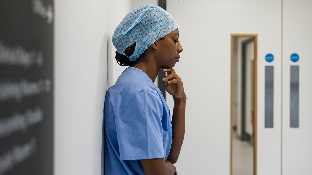 An NHS hospital worker wearing scrubs and a hair net stands against a wall inside a hospital in Newcastle while looking pensive. Stock photo illustration.