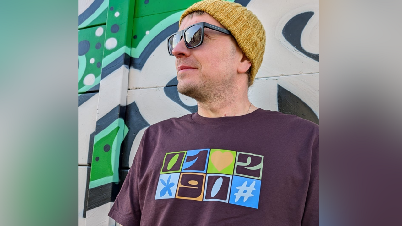 Tim stands next to a green mural behind him and is looking to the left. He has a beanie hat on, shades and a purple T'shirt with the area code on.