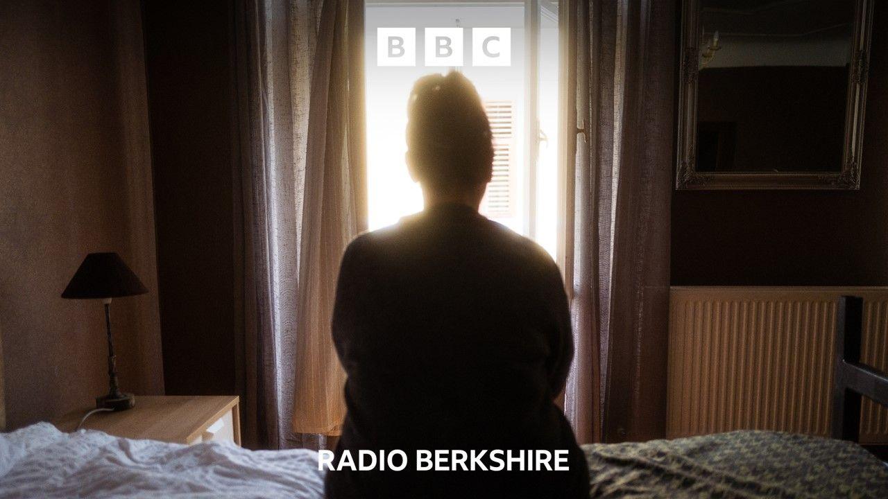 A woman sits on a bed, silhouetted against the light  from a window. Her back is to the camera.