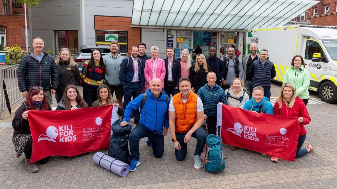 More than 20 people posing for the camera outside a building. Mike Weaver (centre right) has one knee on the ground and is wearing an orange top. Two identical red and white banners, including the words Kili for kids, are being held by other people. 