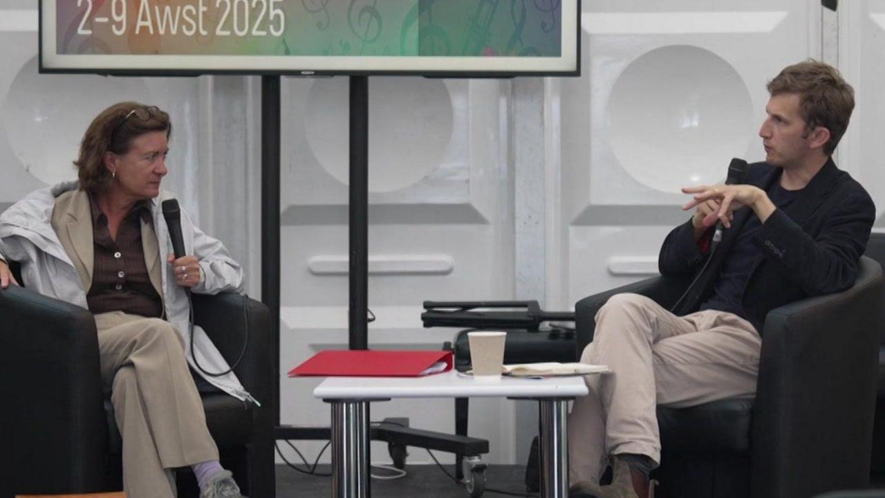 Eluned Morgan and Owain Williams on stage at the National Eisteddfod. They are sat in chairs with a table in the middle of them, with a coffee cup and a binder.