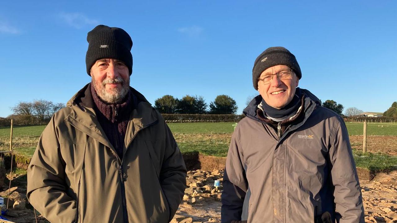 Chris and Bill smile as they pose at the site with rocks from an excavated pit in the green famrland behind them. They both wear balck beanie hats and dark coats on a sunny frosty day.