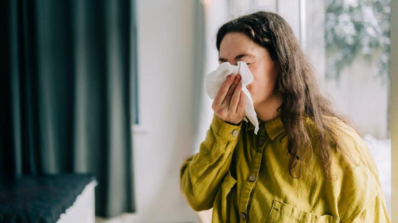 A woman with brown long frizzy hair wearing a lime green shirt holding a tissue up to her face and blowing her nose. The background is blurred but she is inside.