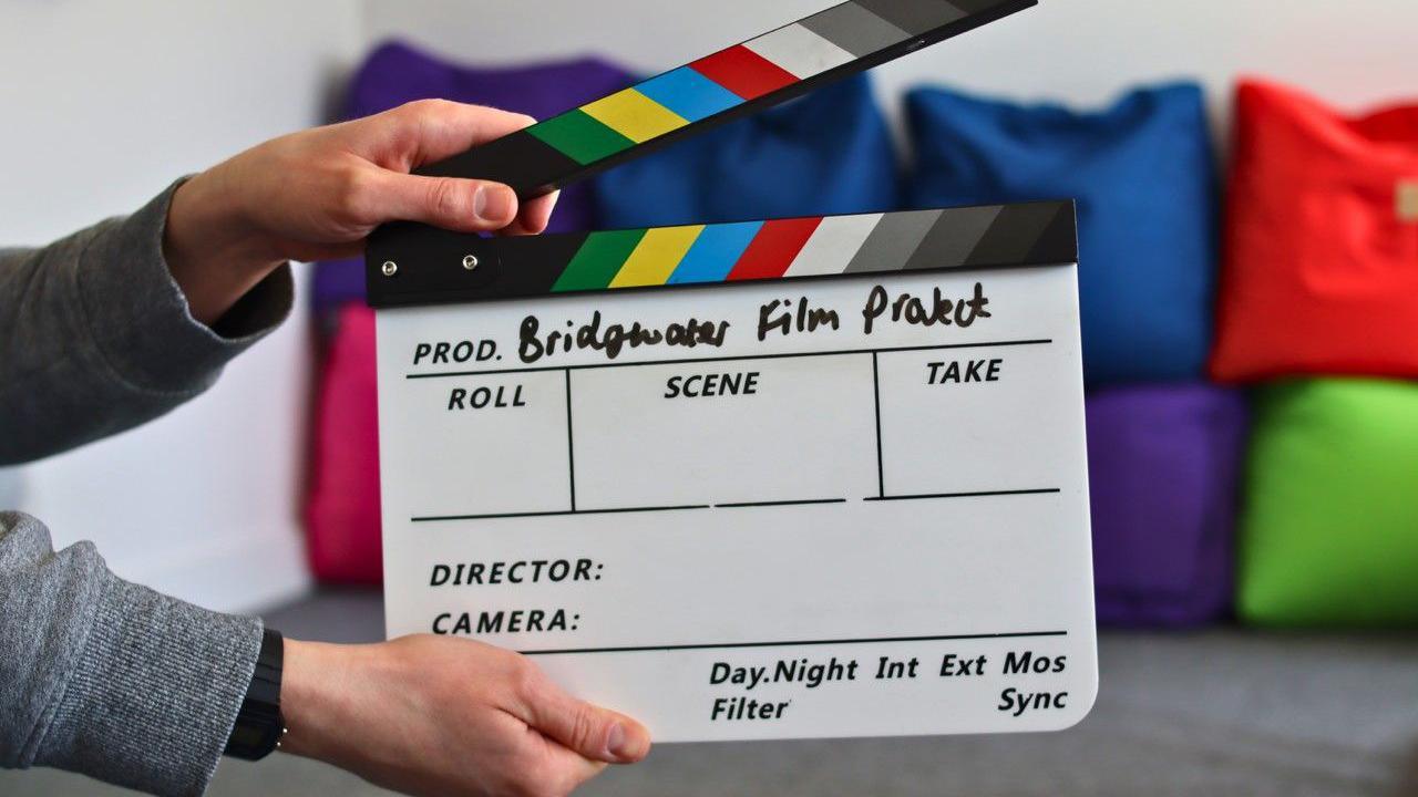 A person, out of view, holds a film slate with two hands. There is a pile of what look like multicoloured bean bags in the background. 