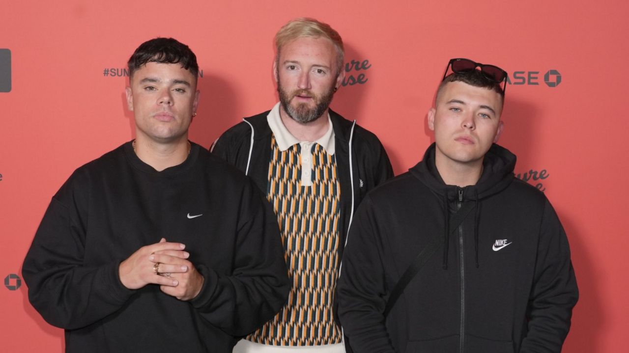 Kneecap members Liam Og O Hannaigh (Mo Chara), JJ O'Dochartaigh (DJ Provai), and Naoise O Caireallain (Moglai Bap) attending the UK premiere of Kneecap, at the Picturehouse Central Cinema, London. The group are wearing black Nike jumpers and are standing in front of a red background.