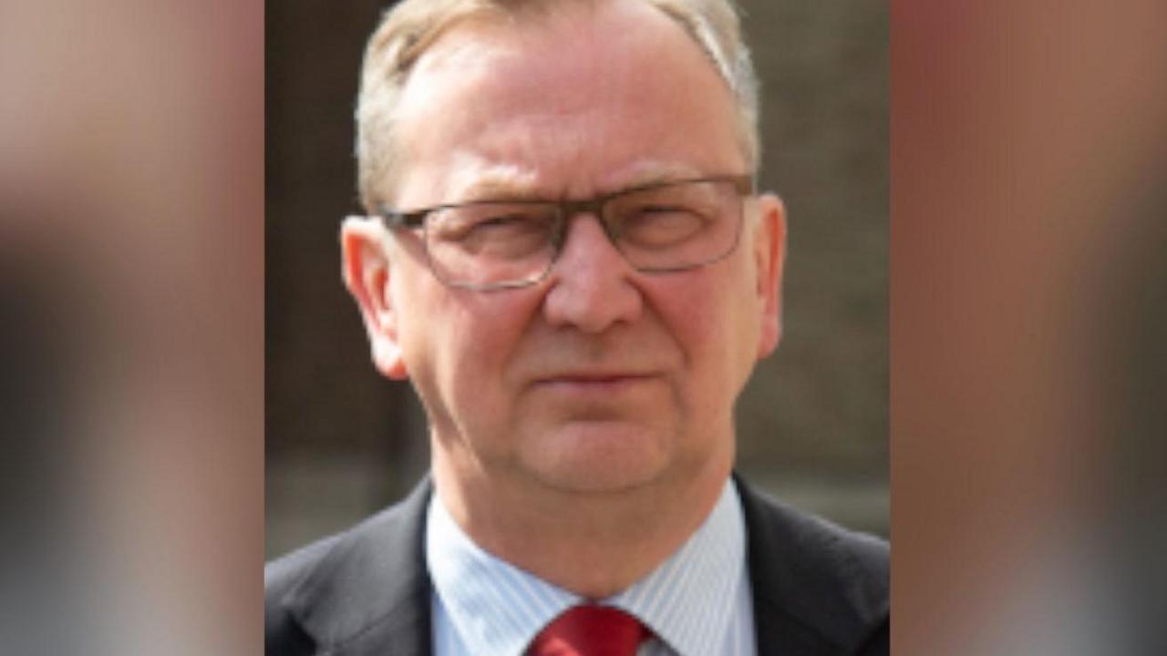 A head and shoulders shot of councillor Fabian Breckels, who is is squinting as he looks at the camera. He has short grey hair and wears rectangular, thin-rimmed glasses. He is wearing a black suit jacket, white and blue pin-striped shirt and a red tie.