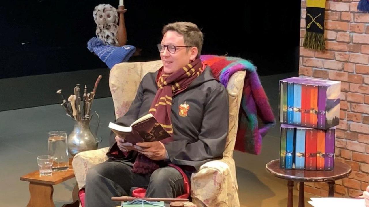 Joe Edwards, who has dark hair and wearing a Harry Potter fancy dress costume sitting in a chair on a theatre stage holding a copy of a book. to his left there is a pot of wands on a table. To his right there is copies of the wizarding saga. 