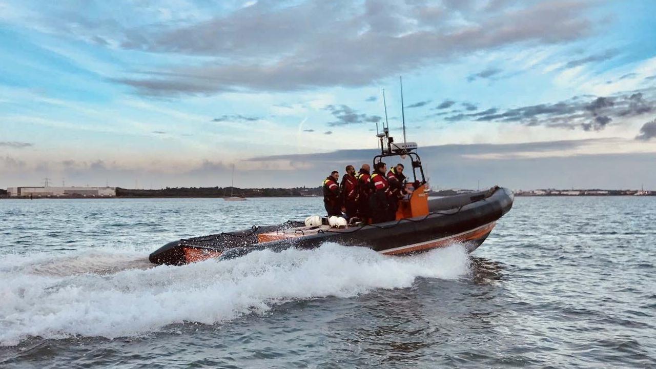 A lifeboat crew stand together on a boat, which looks to be moving at sea. They are all wearing red reflective clothing.